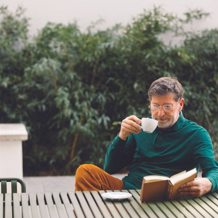 Elderly man sitting at an outdoor table reading a book in a peaceful garden setting