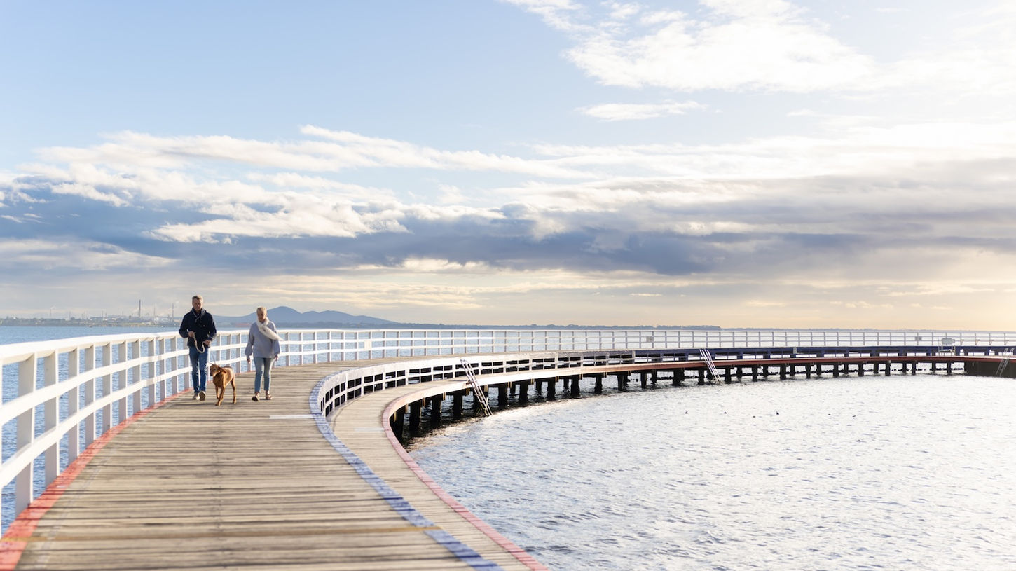 Family walking along a curved wooden boardwalk pier stretching into a calm bay at sunset